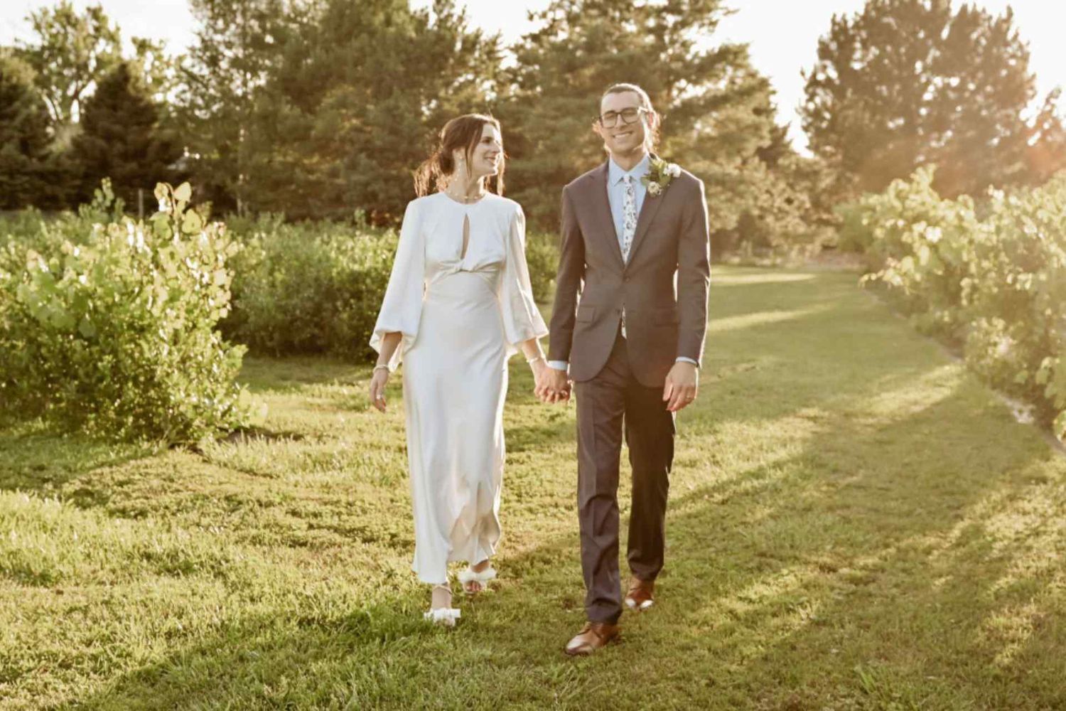 Wedding couple walking along a sunlit path through green scenery.