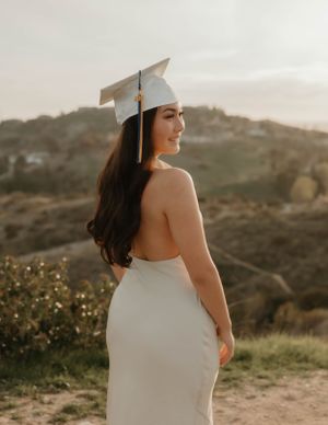 Graduate in white dress and cap poses against scenic hillside backdrop at sunset for graduation and senior photos in Orange County