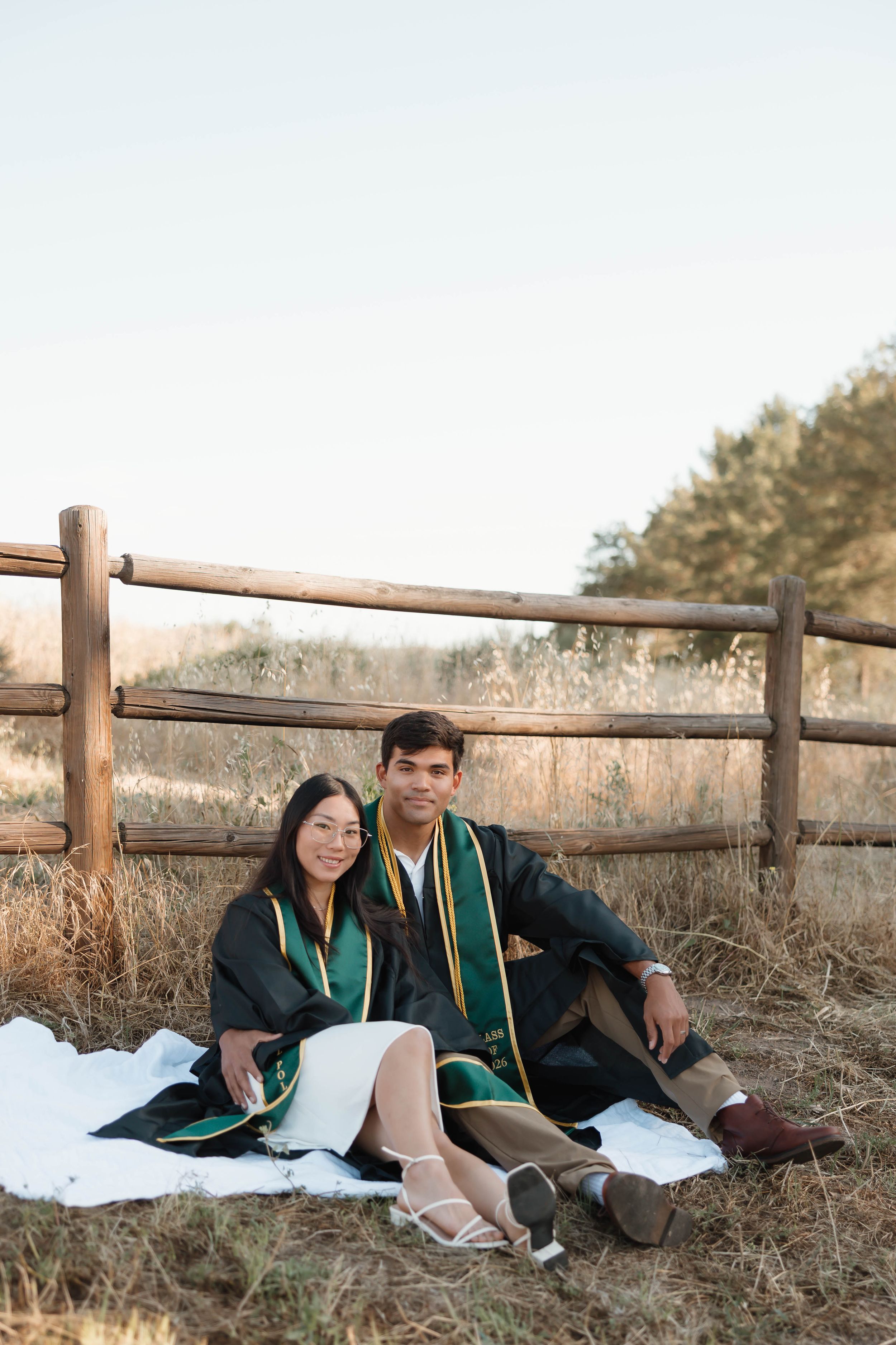 Two graduates in black and green gowns sit on a blanket by a wooden fence in a golden field.