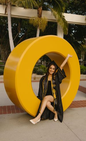 Graduate in academic regalia sitting inside large yellow letter O sculpture outdoors.