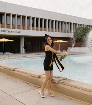 Graduate posing with diploma near fountain outside Brotman Hall building.