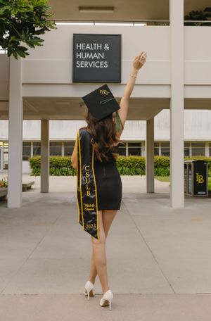 Graduate in black dress and academic regalia poses victoriously under Health Services sign.