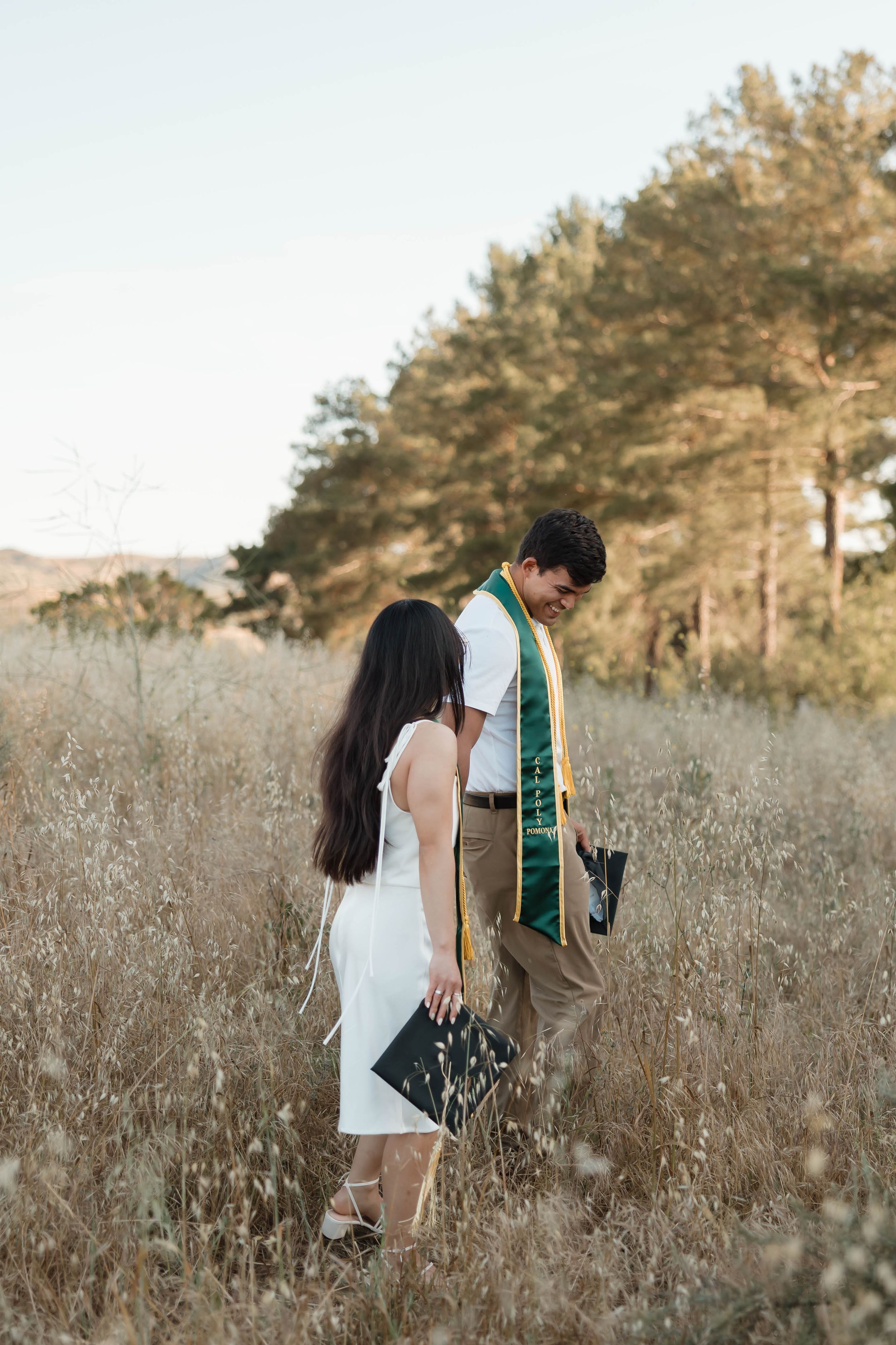 A graduate in green sash and partner in white dress stand in golden field with pine trees at sunset.