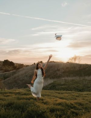 A figure in a flowing white dress leaps joyfully in a sunset field while tossing a graduation cap into the air for graduation and senior pho
