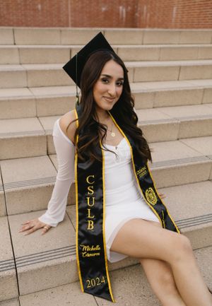 Graduate in white dress poses on steps wearing CSU graduation sash.