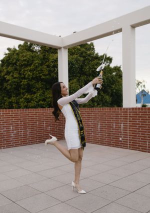 Graduate in white dress playfully poses with graduation cap and sash outdoors.