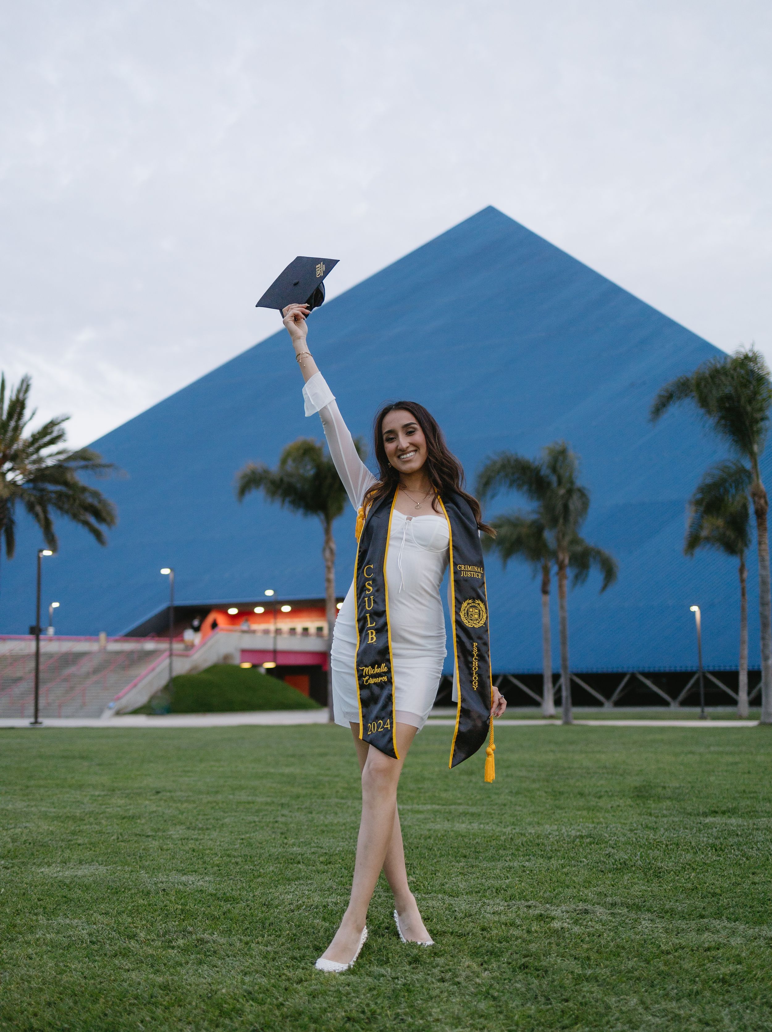 Graduate celebrating with raised cap in front of blue pyramid-shaped building and palm trees on grassy lawn.