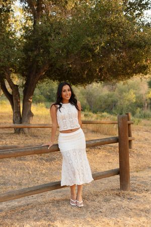 A person in a white two-piece lace outfit stands by a wooden fence under a tree for graduation and senior photos in Orange County