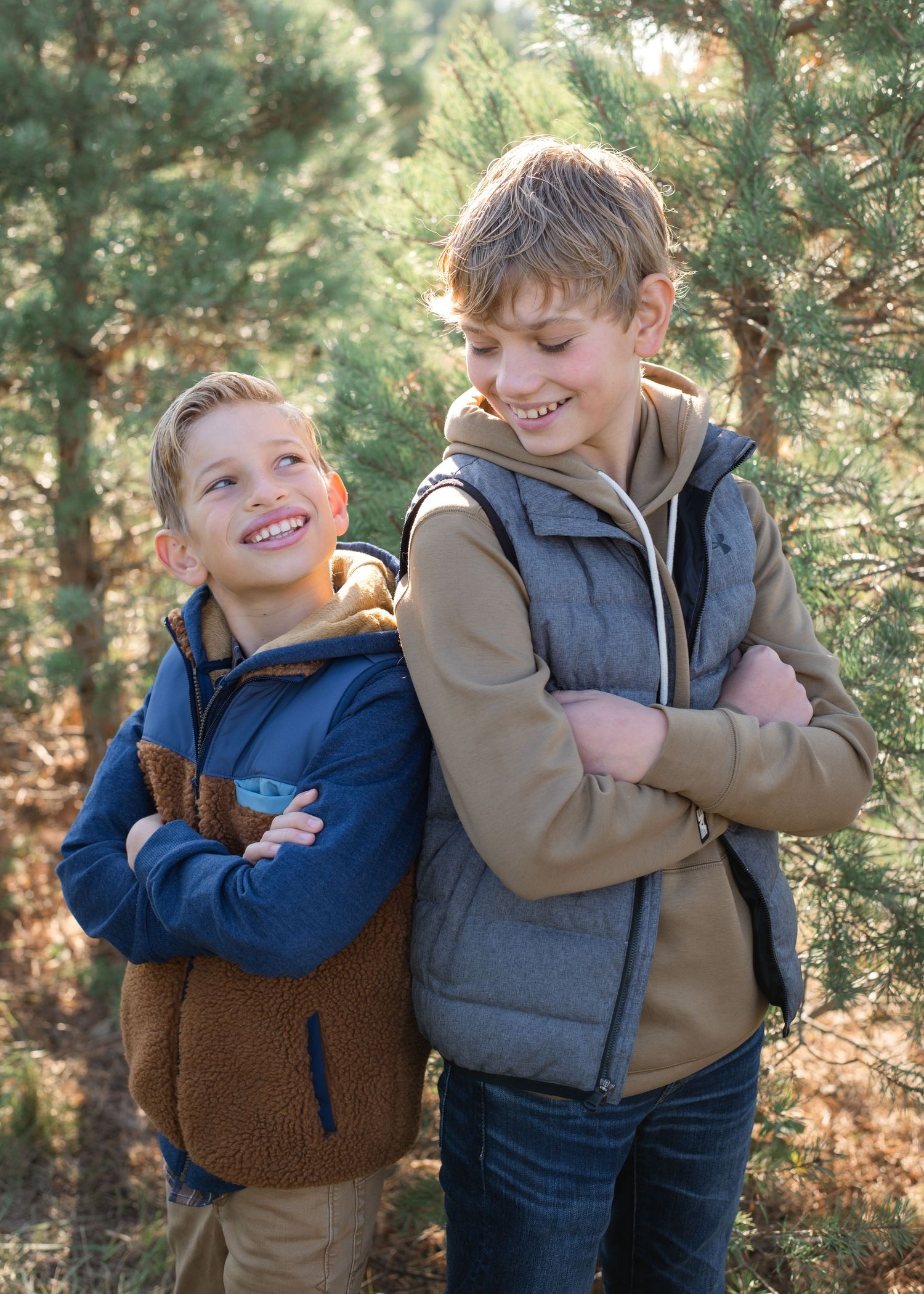 Two young people in casual winter wear smile while standing among pine trees.