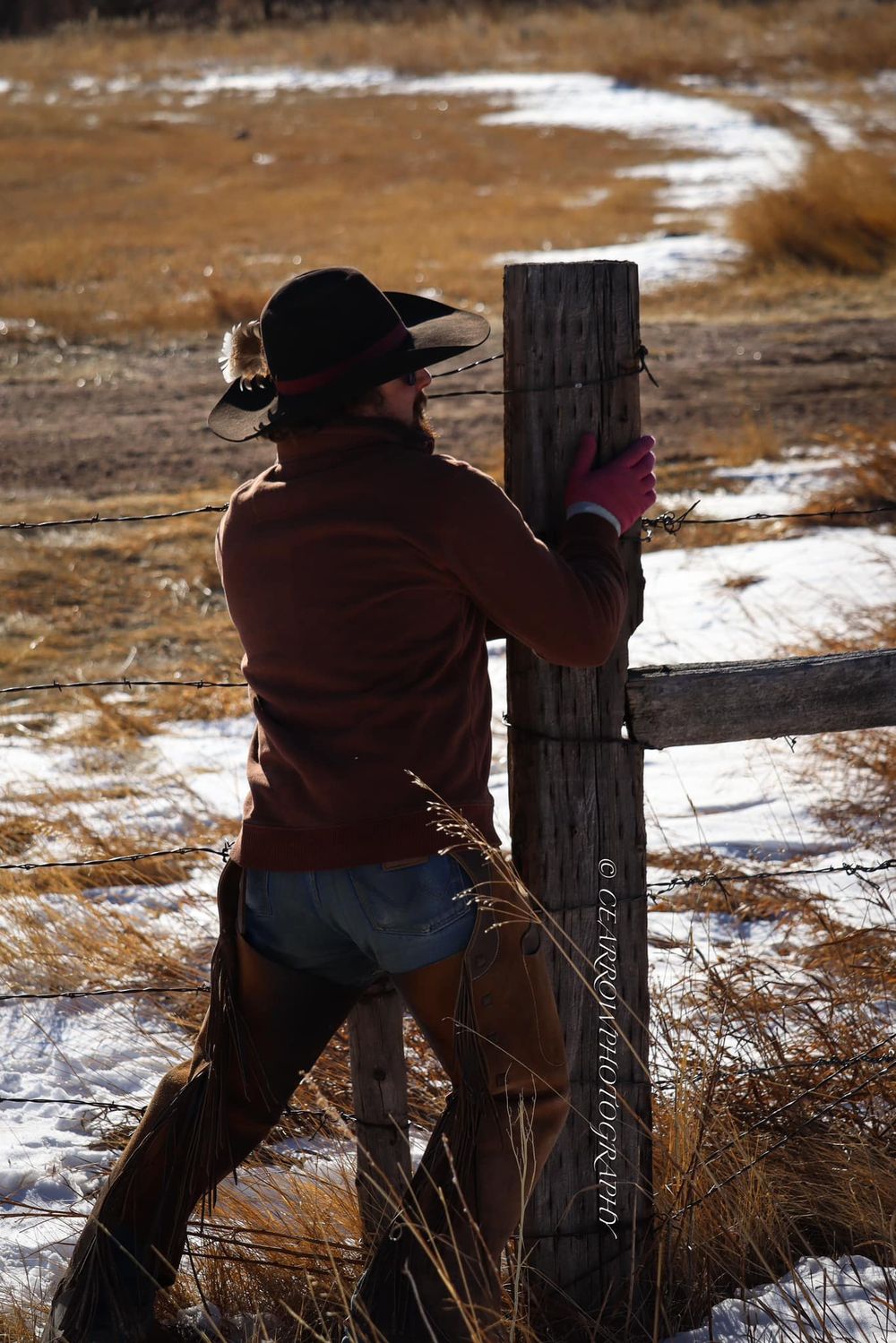 A ranch worker fixes a wooden fence post in winter with snowy ground visible.
