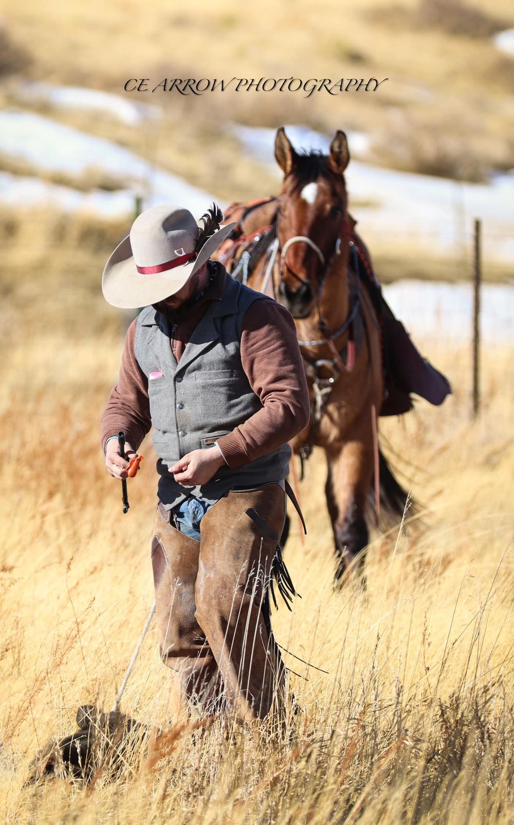 Cowboy walking through tall grass leading brown horse in western landscape.