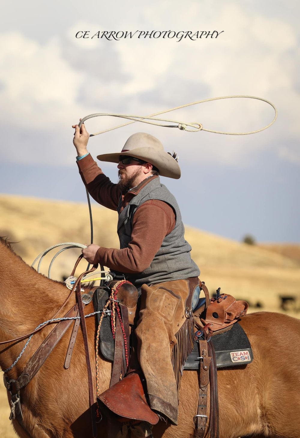 A cowboy on horseback swings a lasso against a cloudy sky in western ranch country.