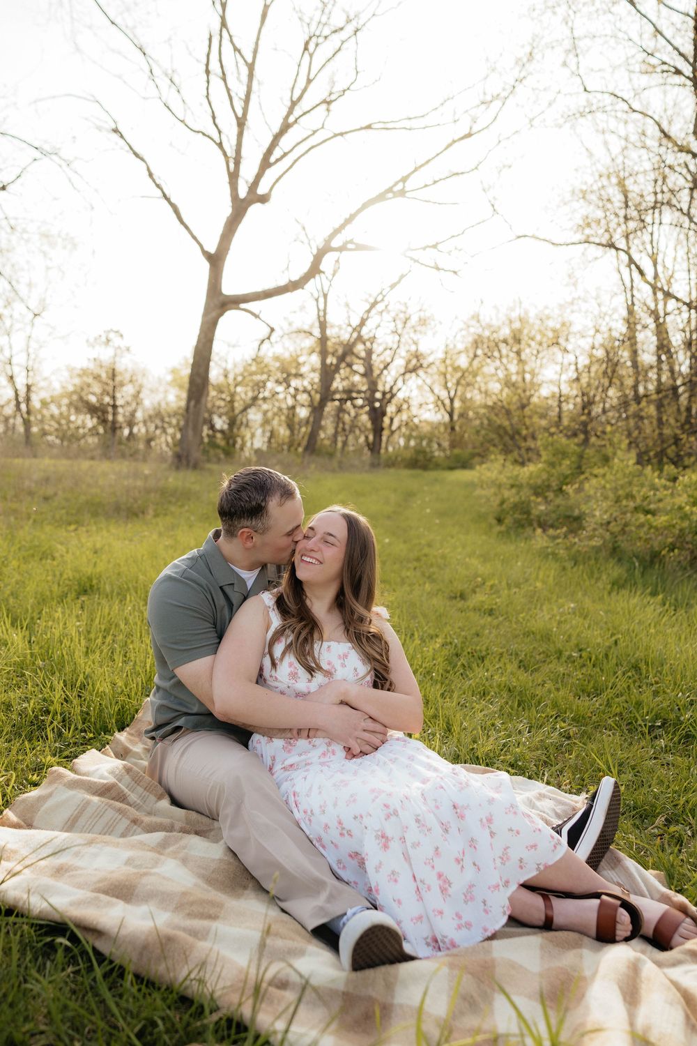 Couple sitting on a blanket in a sunny meadow, man kissing woman's cheek as she smiles in a floral dress.