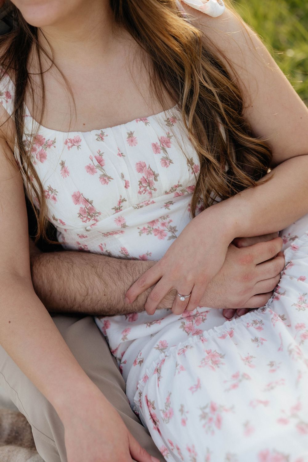 Couple sitting together outdoors, woman wearing white floral dress with pink flowers, man's arm wrapped around her waist.