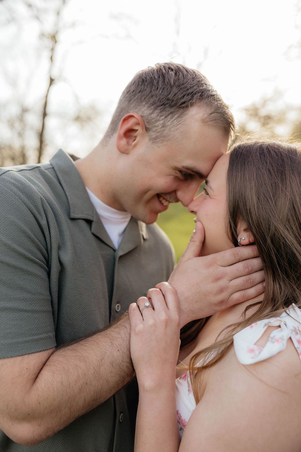 A couple shares a tender forehead-to-forehead moment outdoors, smiling closely as the man gently cups the womans face.