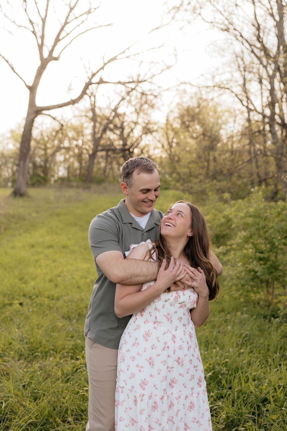Couple embracing in a sunlit green meadow, woman in floral dress laughing joyfully as man hugs her from behind.