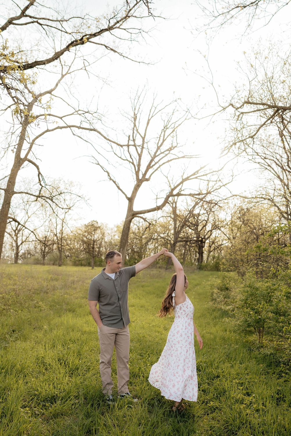 A couple dances outdoors in a green meadow, man spinning woman in white floral dress among bare trees at golden hour.