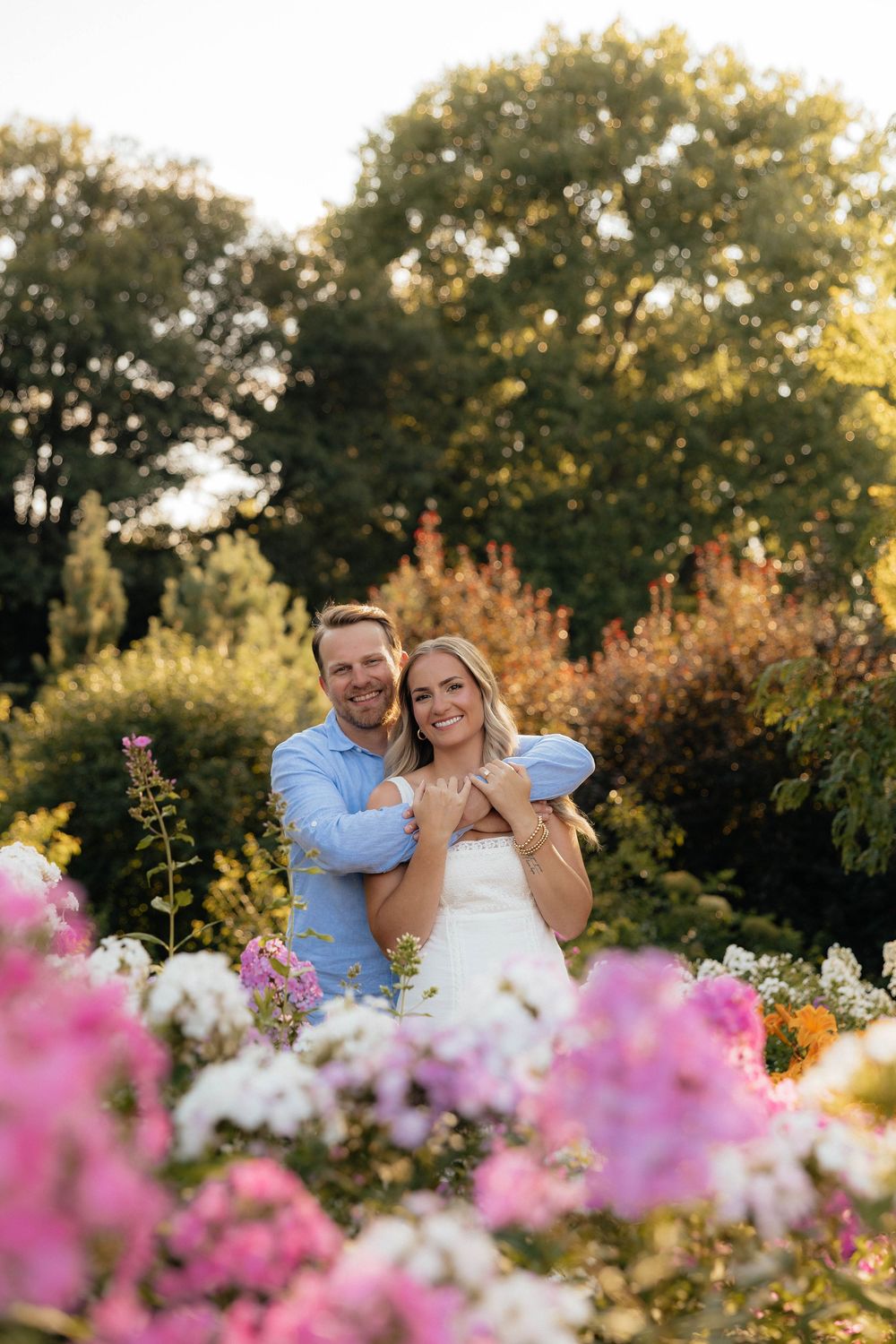 Couple embracing in a colorful garden, man in blue shirt hugging smiling woman in white dress, surrounded by pink and white flowers.