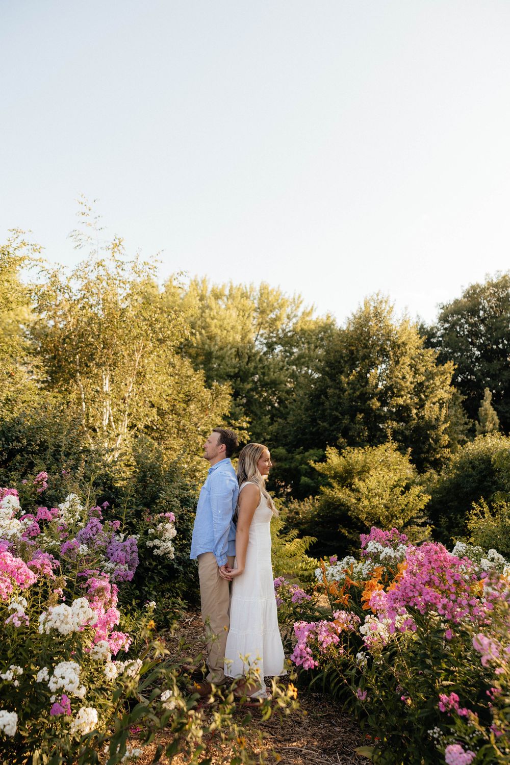 Couple standing back-to-back in a lush garden surrounded by pink and white flowers on a sunny day.