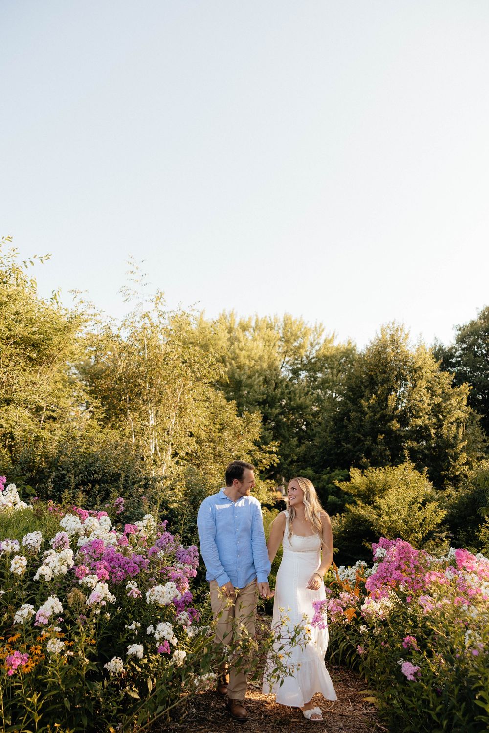 Couple holding hands in a lush garden surrounded by pink and white flowers during golden hour.
