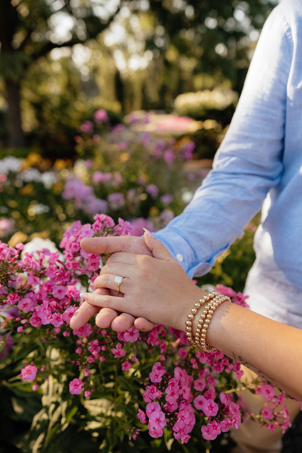 Two hands clasped together over pink flowers, featuring a diamond engagement ring and gold beaded bracelet in a garden setting.
