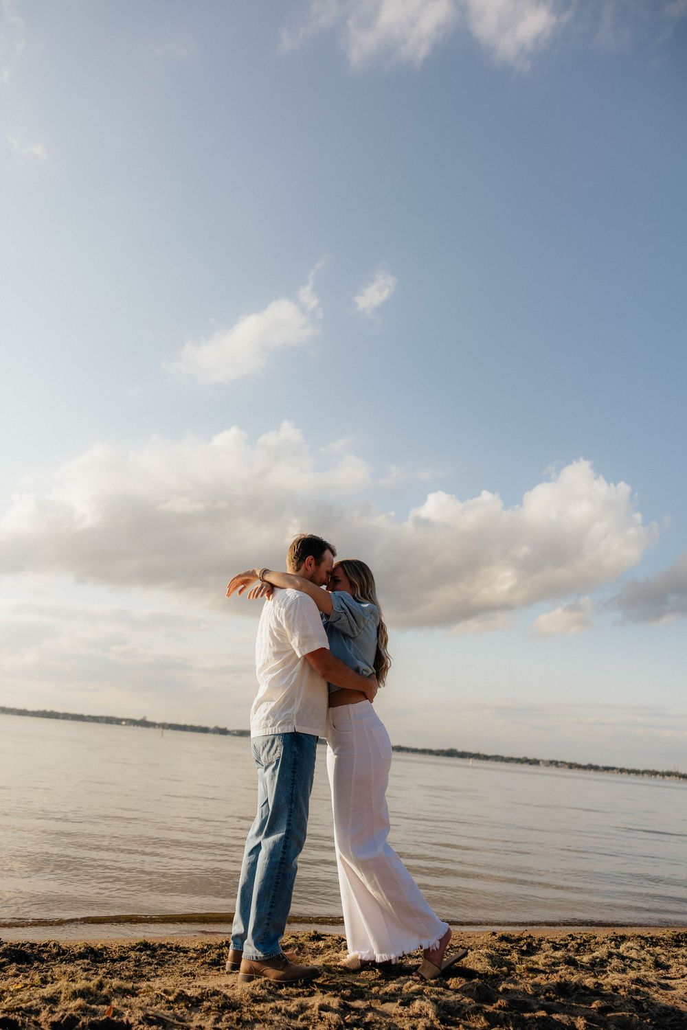 Couple embracing and kissing on a sandy beach shore with a blue sky and fluffy clouds in the background.