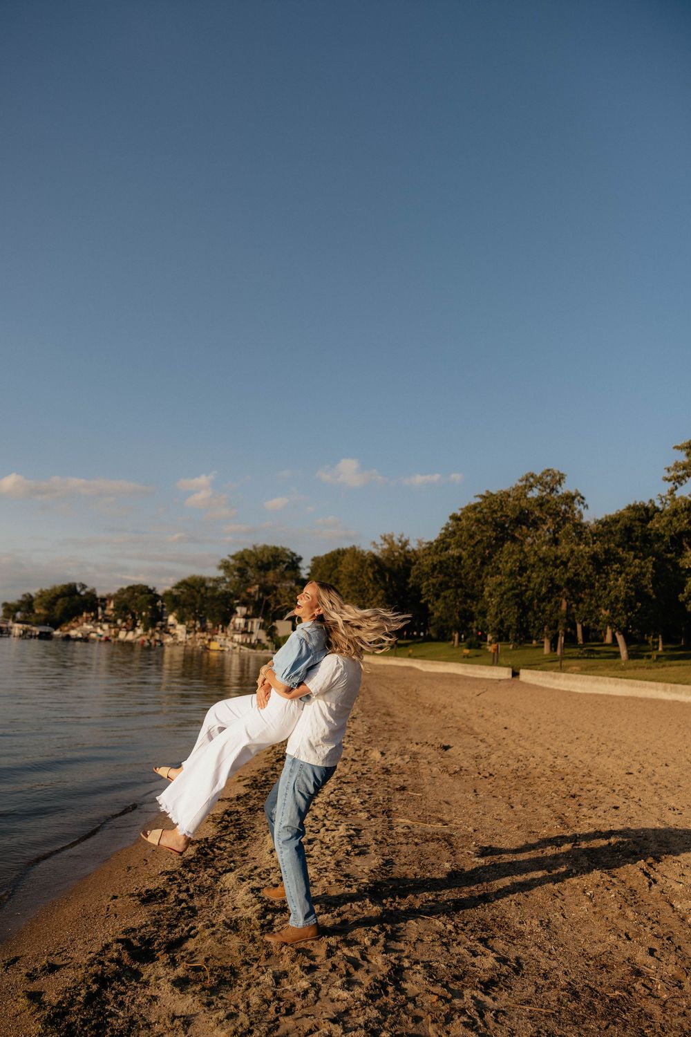 Couple embracing on sandy beach at golden hour, woman lifted in air, trees and calm water in background.