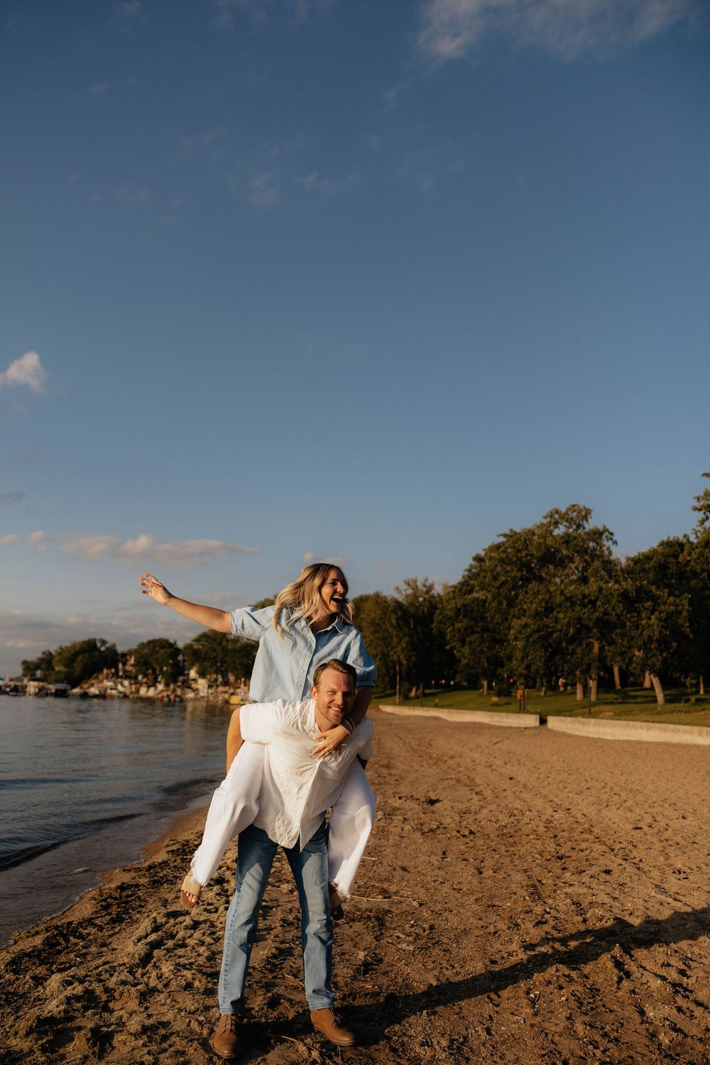 A couple enjoys a piggyback ride on a sandy beach at golden hour, with the woman spreading her arms joyfully.