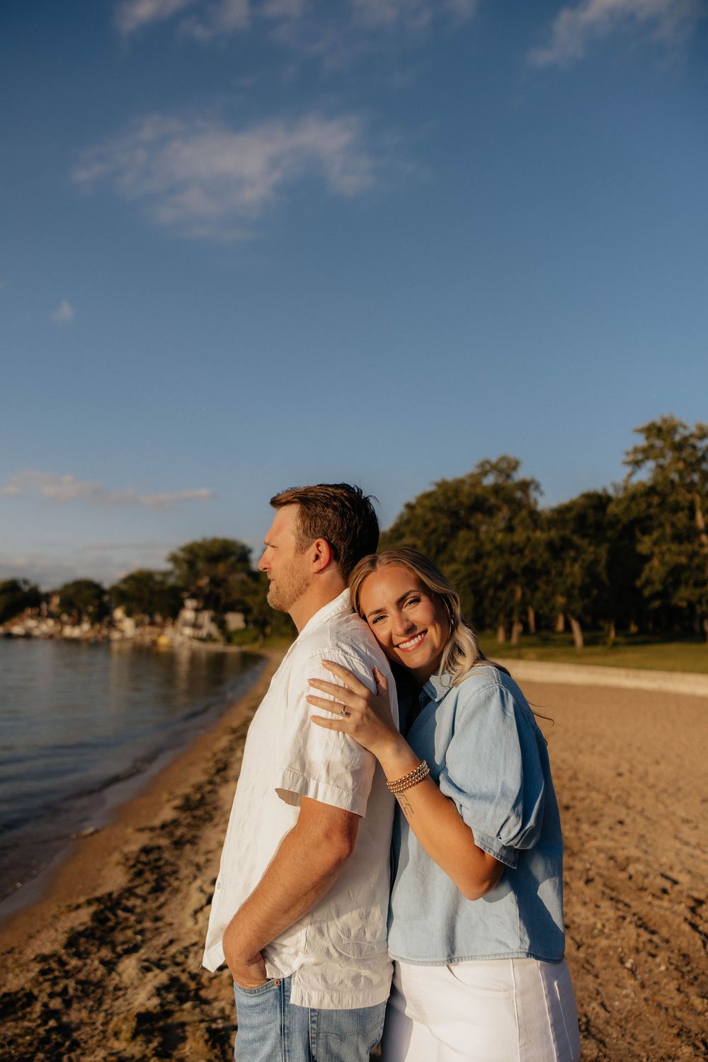 Couple embracing on a sandy beach at golden hour, woman in blue shirt smiling while hugging man in white shirt.