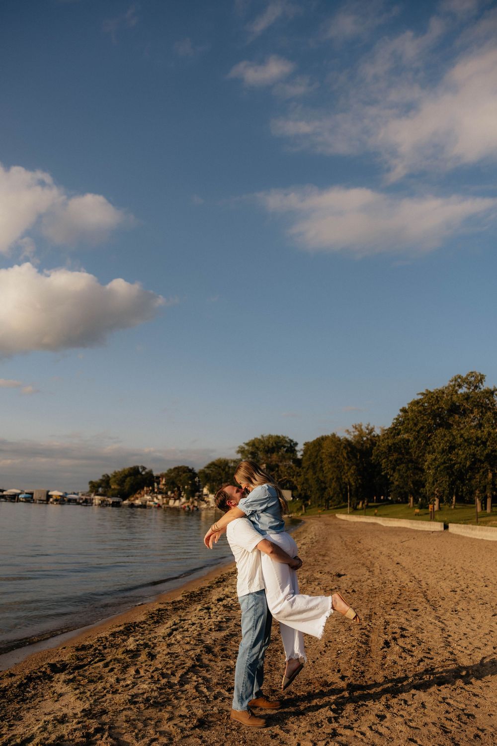 Couple embracing on sandy beach at golden hour with blue sky, trees, and calm water in the background.