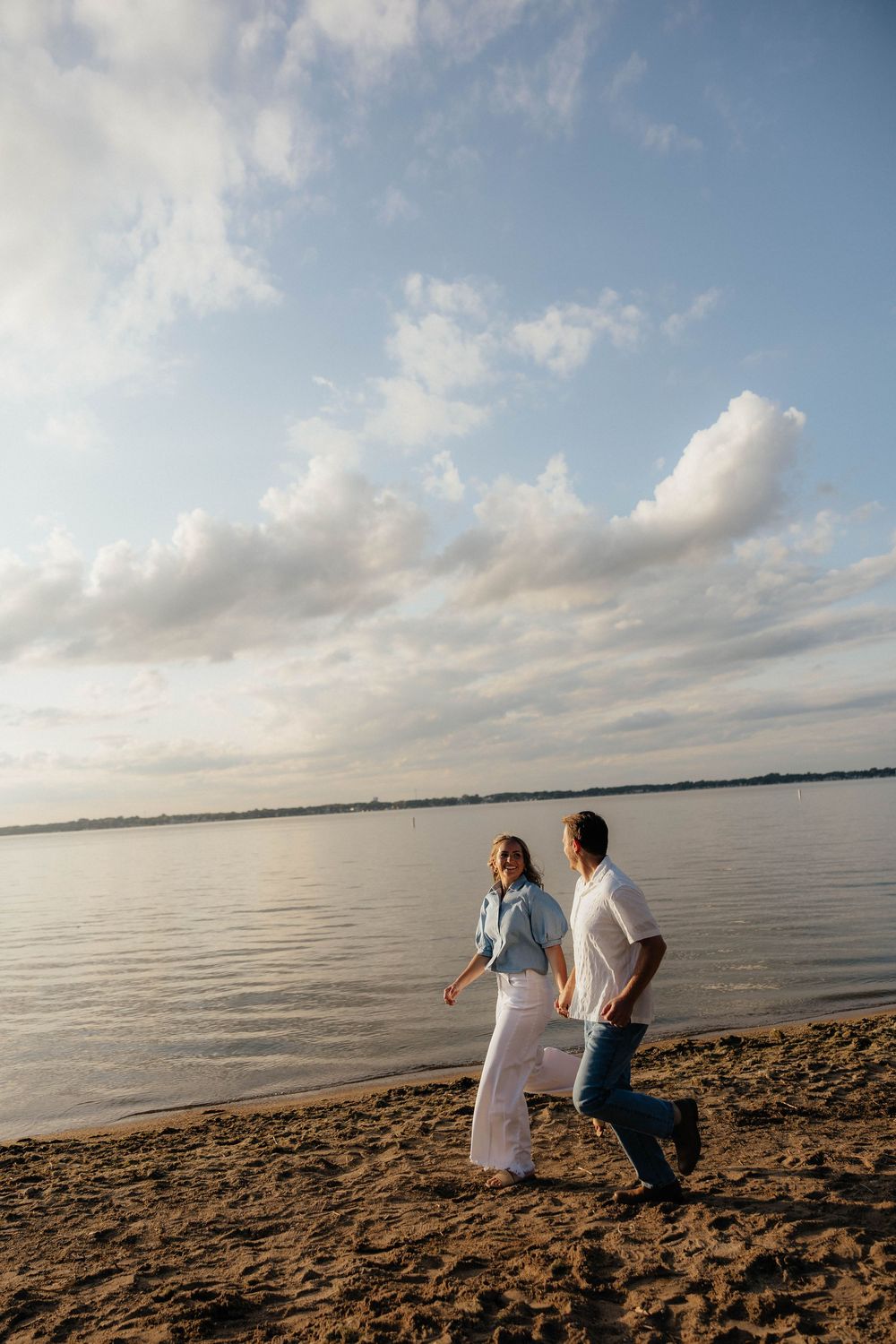 Couple walking hand in hand on a sandy beach shore with calm water and cloudy sky at sunset.