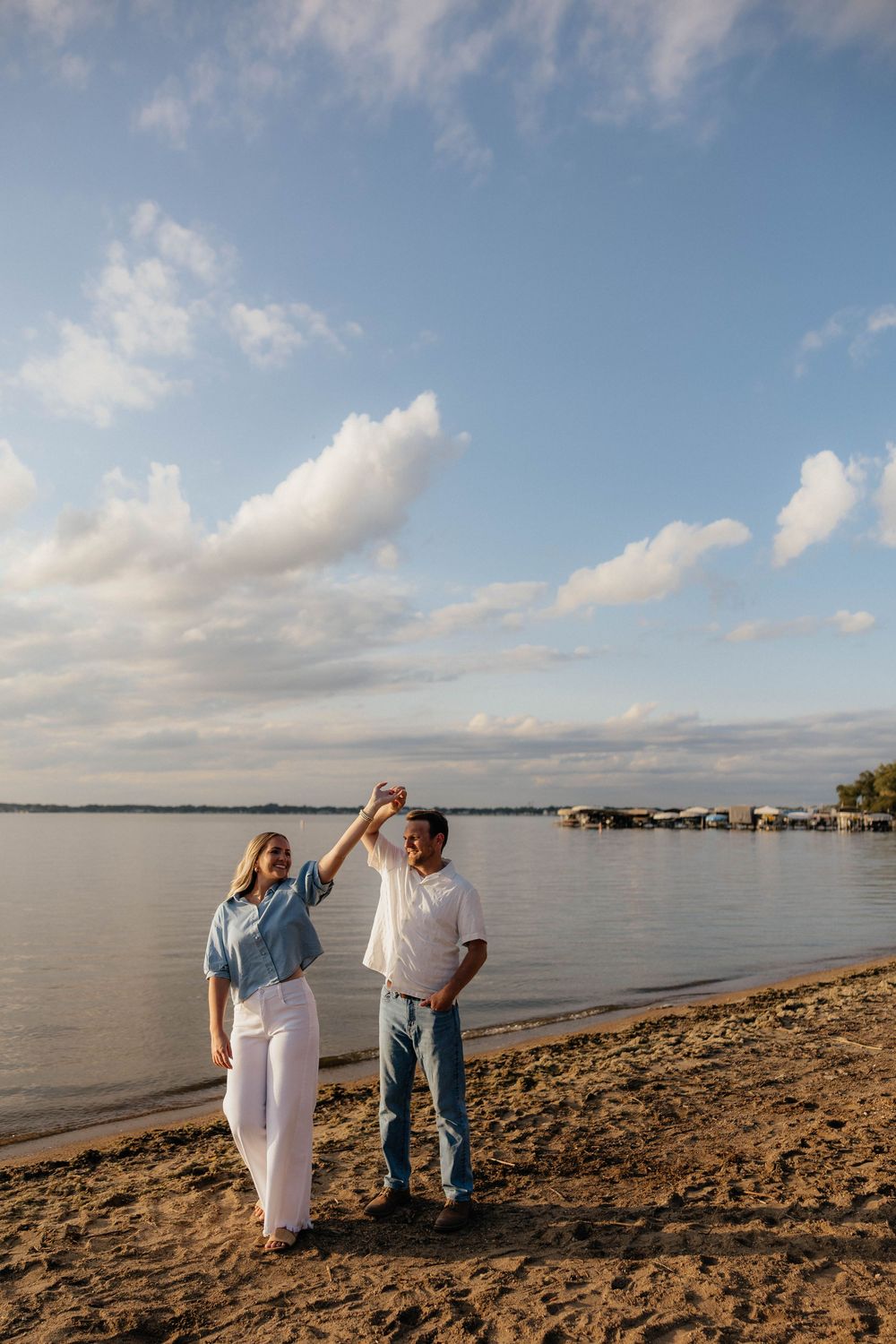 Couple dancing on a sandy beach at sunset, woman in blue shirt and white pants spinning under mans raised hand.