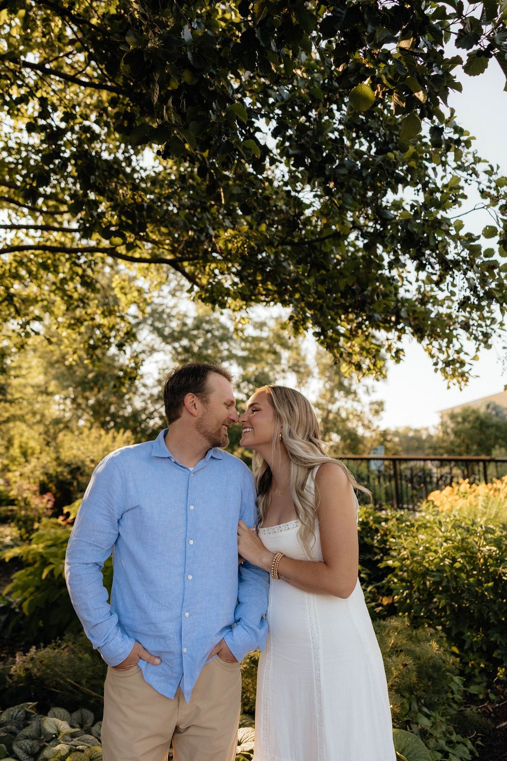 Couple sharing a romantic moment under a lush green tree in a sunlit garden, man in blue shirt and woman in white dress.