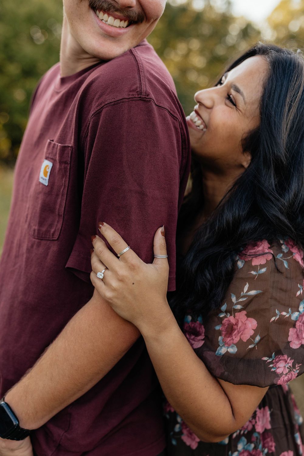 A couple shares joyful laughter during an outdoor engagement photoshoot, with one wearing burgundy and the other in floral print.