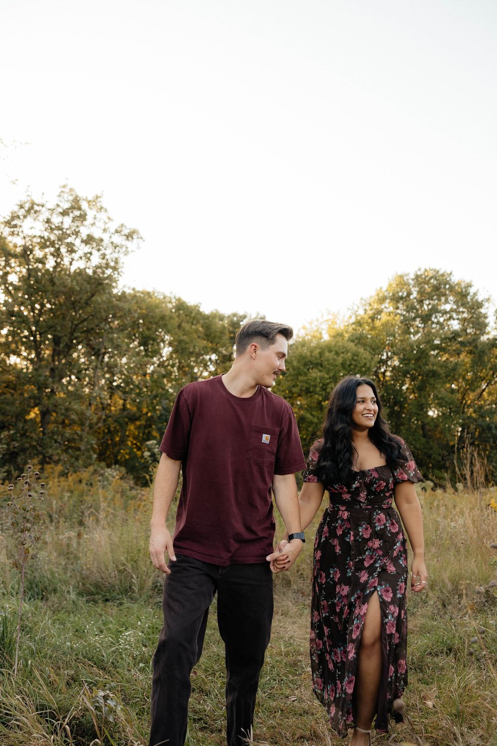 A couple walks hand in hand through a golden field at sunset wearing burgundy and floral outfits.