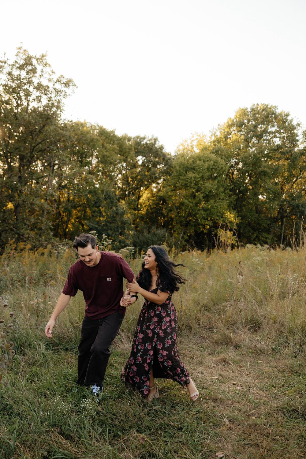 A couple dances and twirls together in a golden field during sunset with trees in the background.