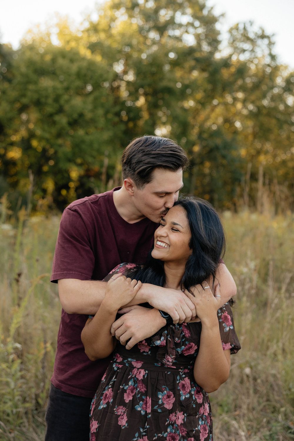 A couple embraces lovingly in a sunlit field with autumn trees in the background during a romantic outdoor photoshoot.