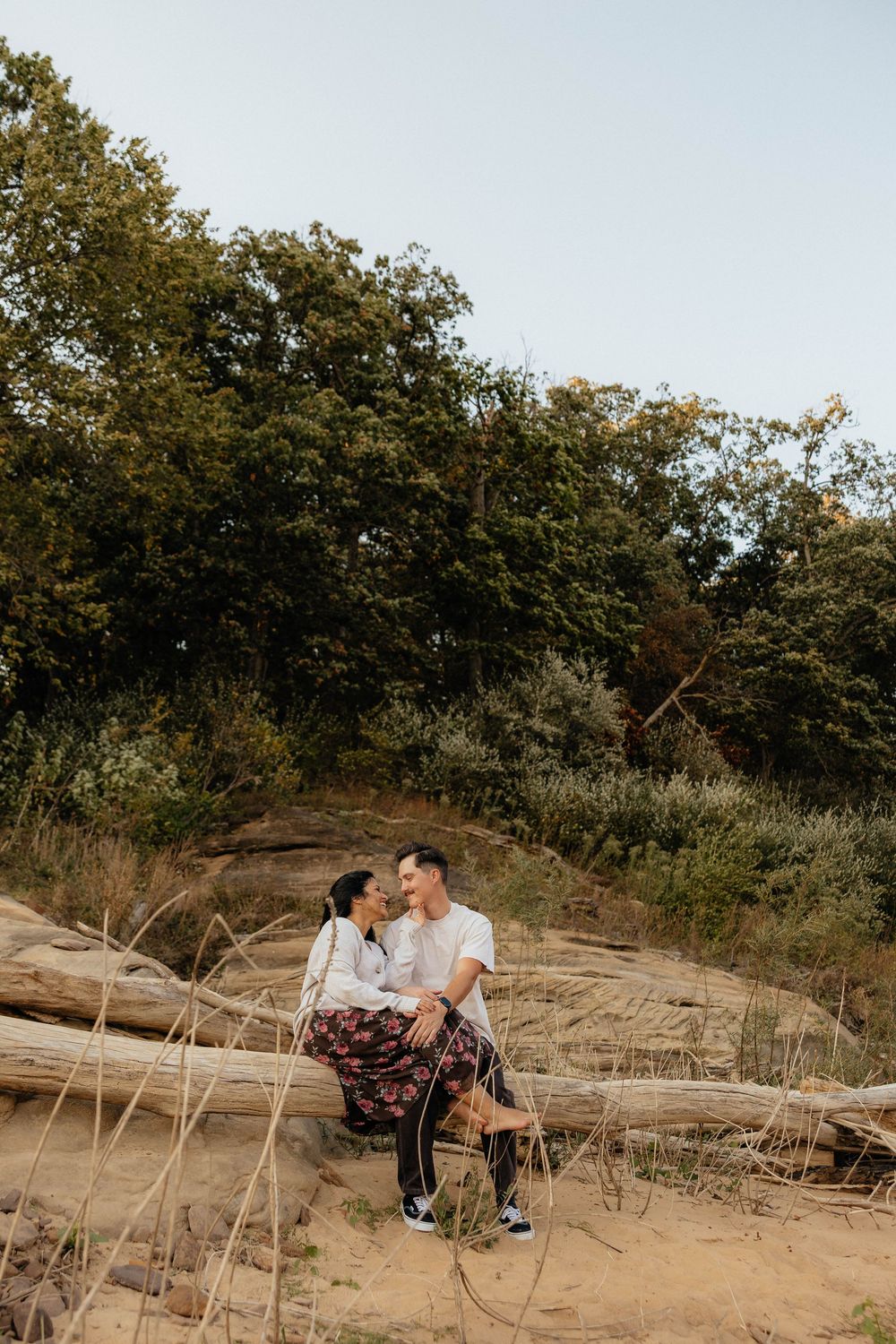 A couple sits together on sandy dunes surrounded by trees and tall grass during a sunset photoshoot.