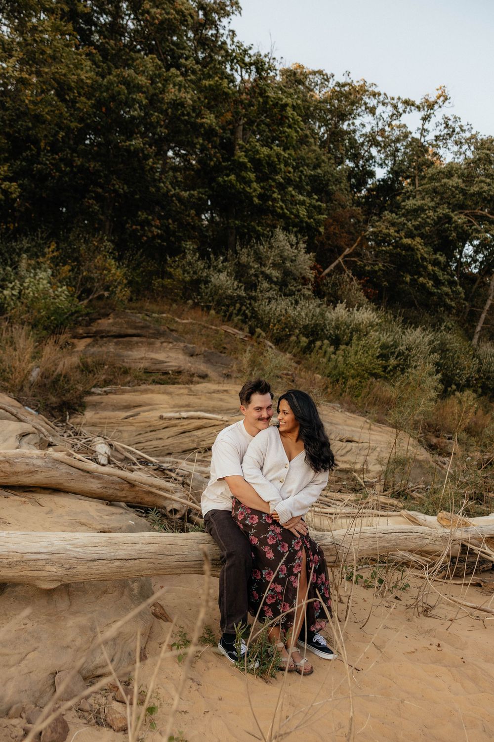 A couple sits together on rocky terrain surrounded by trees and greenery during a romantic outdoor photoshoot.