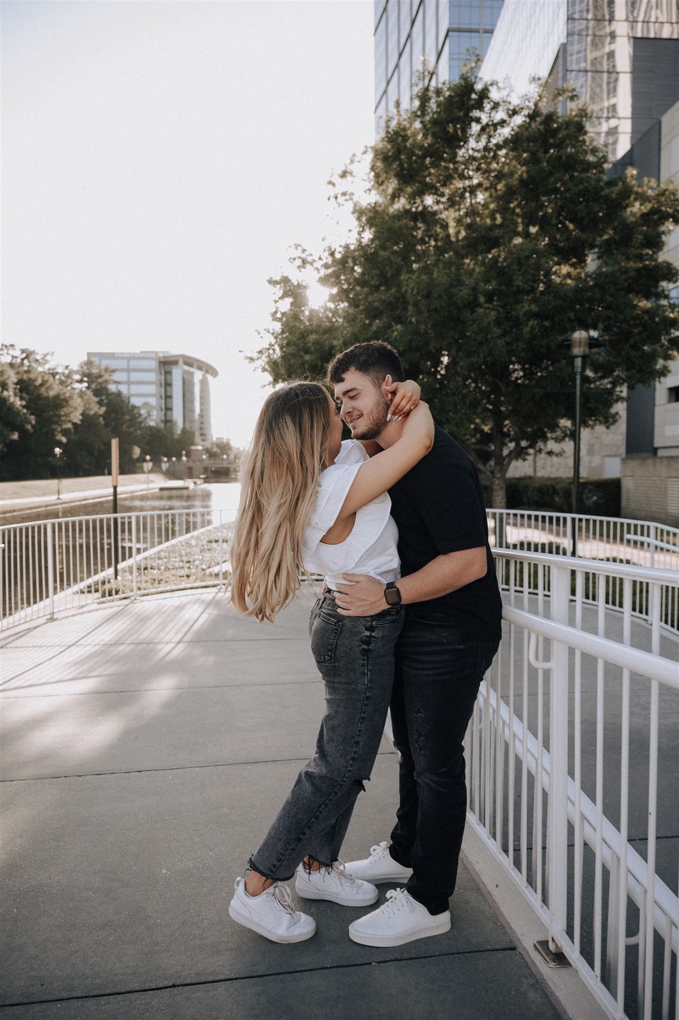 Couple embraces on an urban walkway with white railing and modern buildings in the background during sunset.