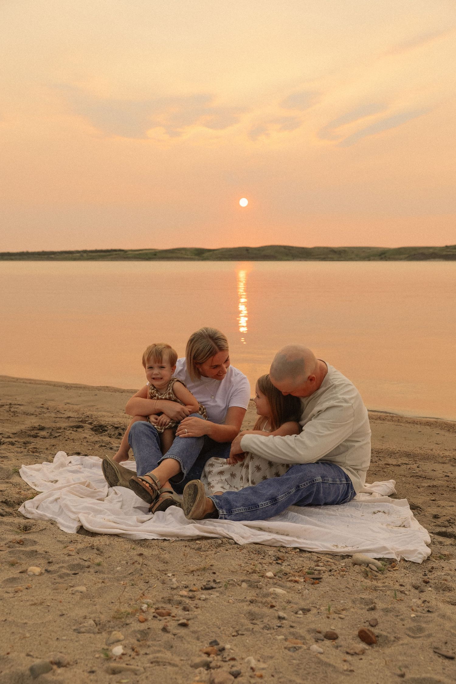 Family lying on a white blanket watching sunset over a lake on a sandy beach.