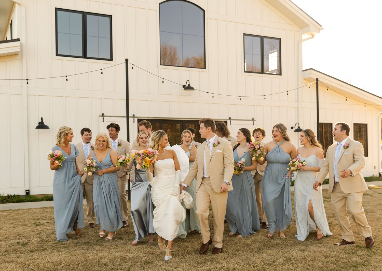 Wedding party in blue and beige attire celebrating outside white venue building.