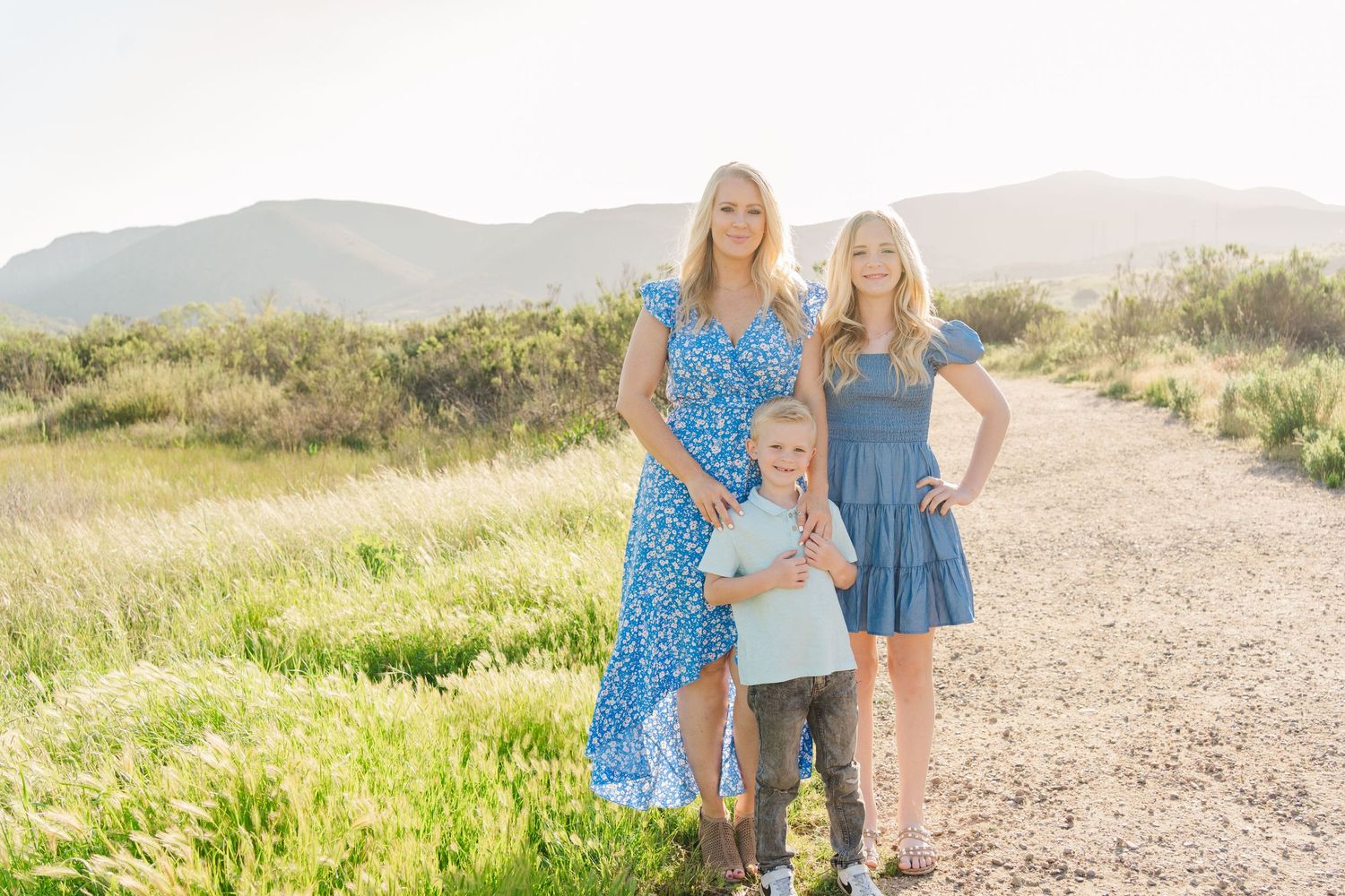 Group wearing coordinating blue dresses stands together on a sunny dirt path with rolling hills in background.