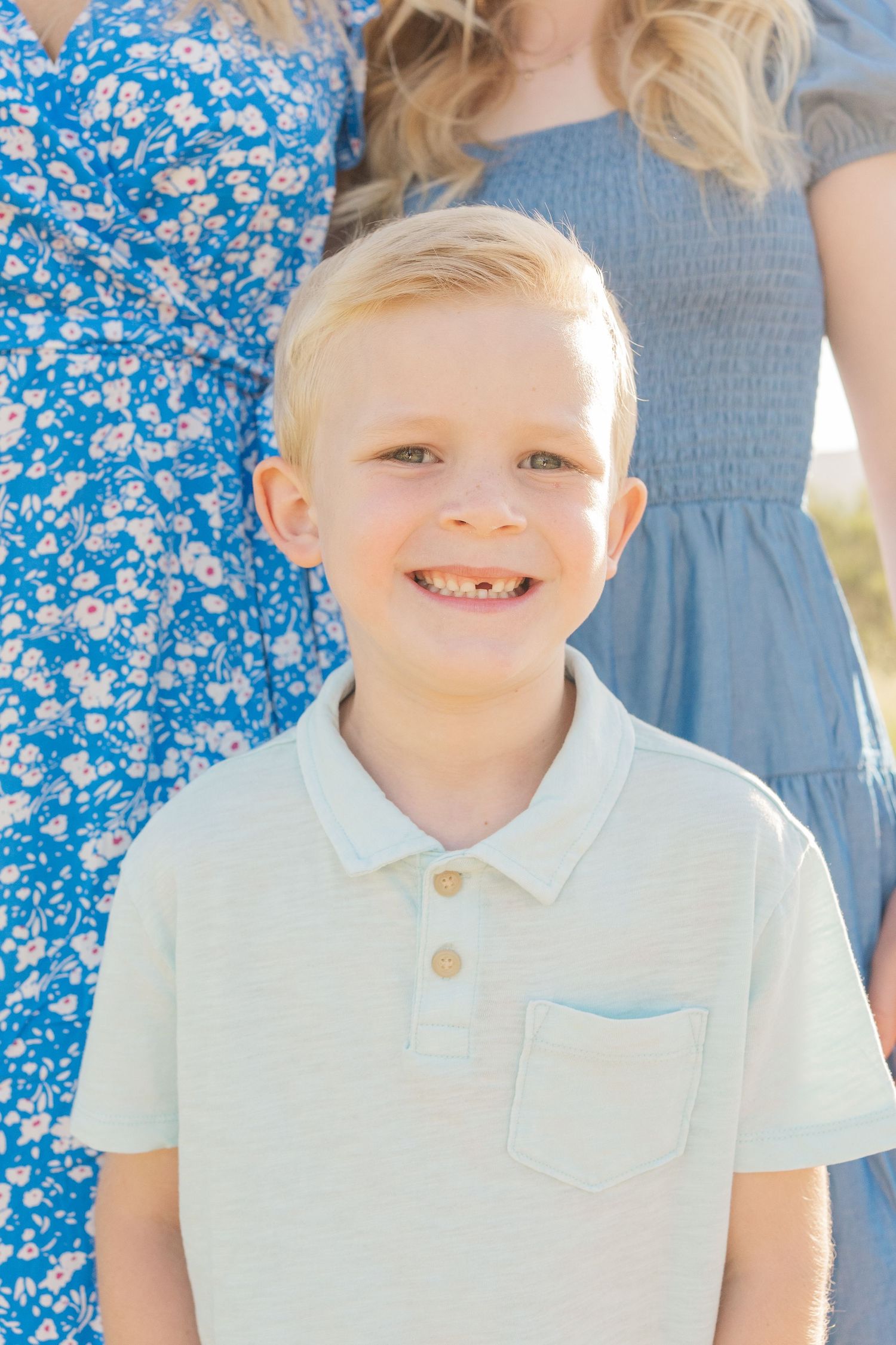 A young child in a white polo shirt smiles outdoors while standing next to someone wearing a blue floral dress.