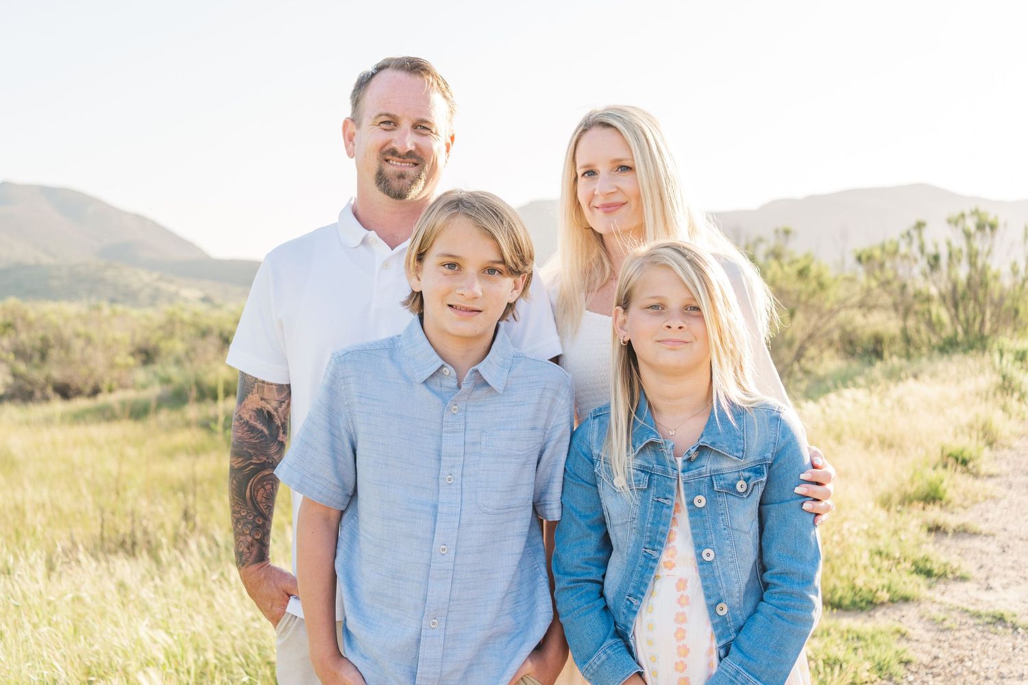 A family of four poses together outdoors on a sunny day with rolling hills and golden grass in the background.
