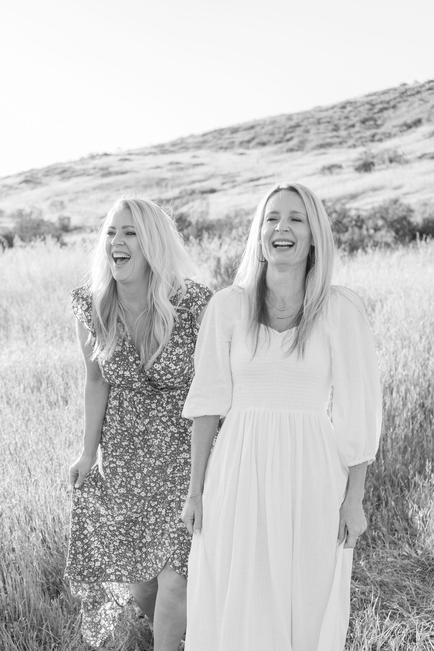 Two friends wearing floral and white dresses laugh together in a sunlit field with hills in the background.