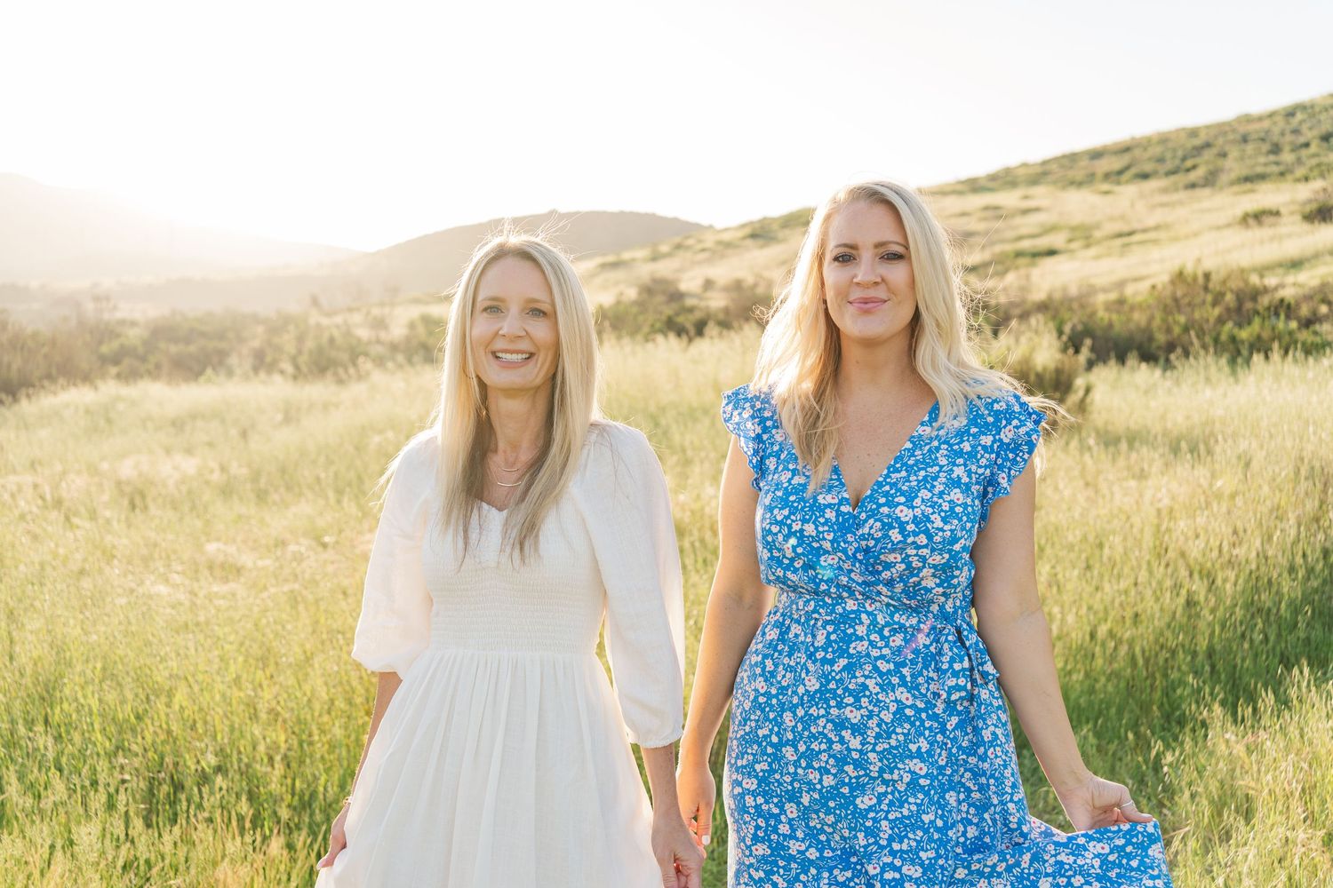 Two people in sundresses standing together in a sunlit meadow with golden grass and rolling hills in the background.