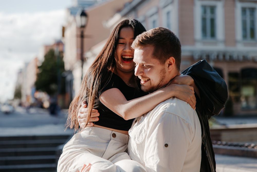 Two people embrace and laugh together during a sunny urban photoshoot.