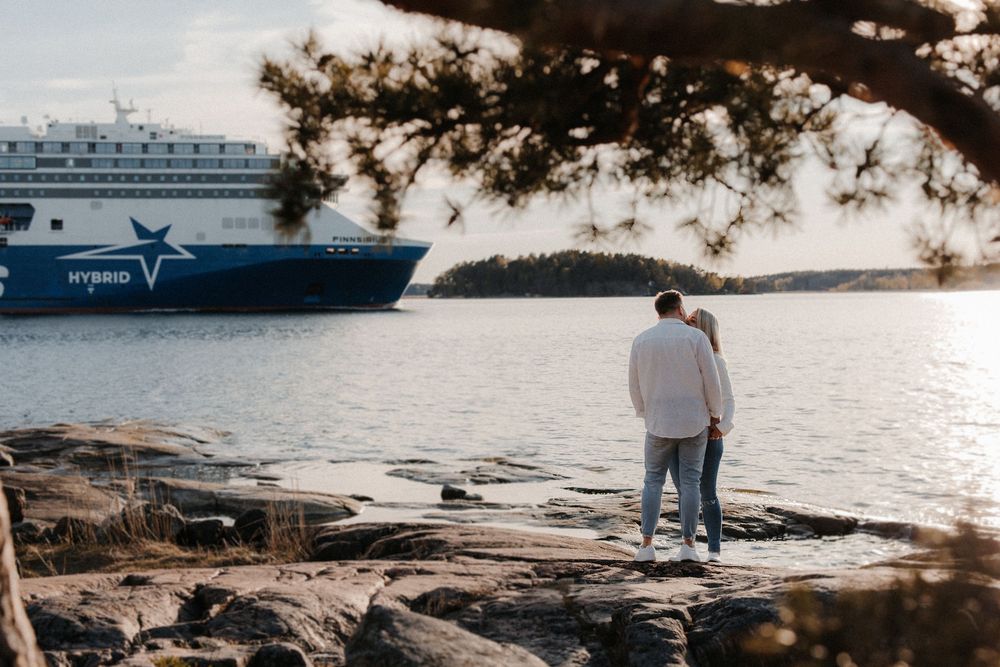 Couple standing on rocky shore watching a Tallink hybrid ferry sail past in calm Nordic waters.