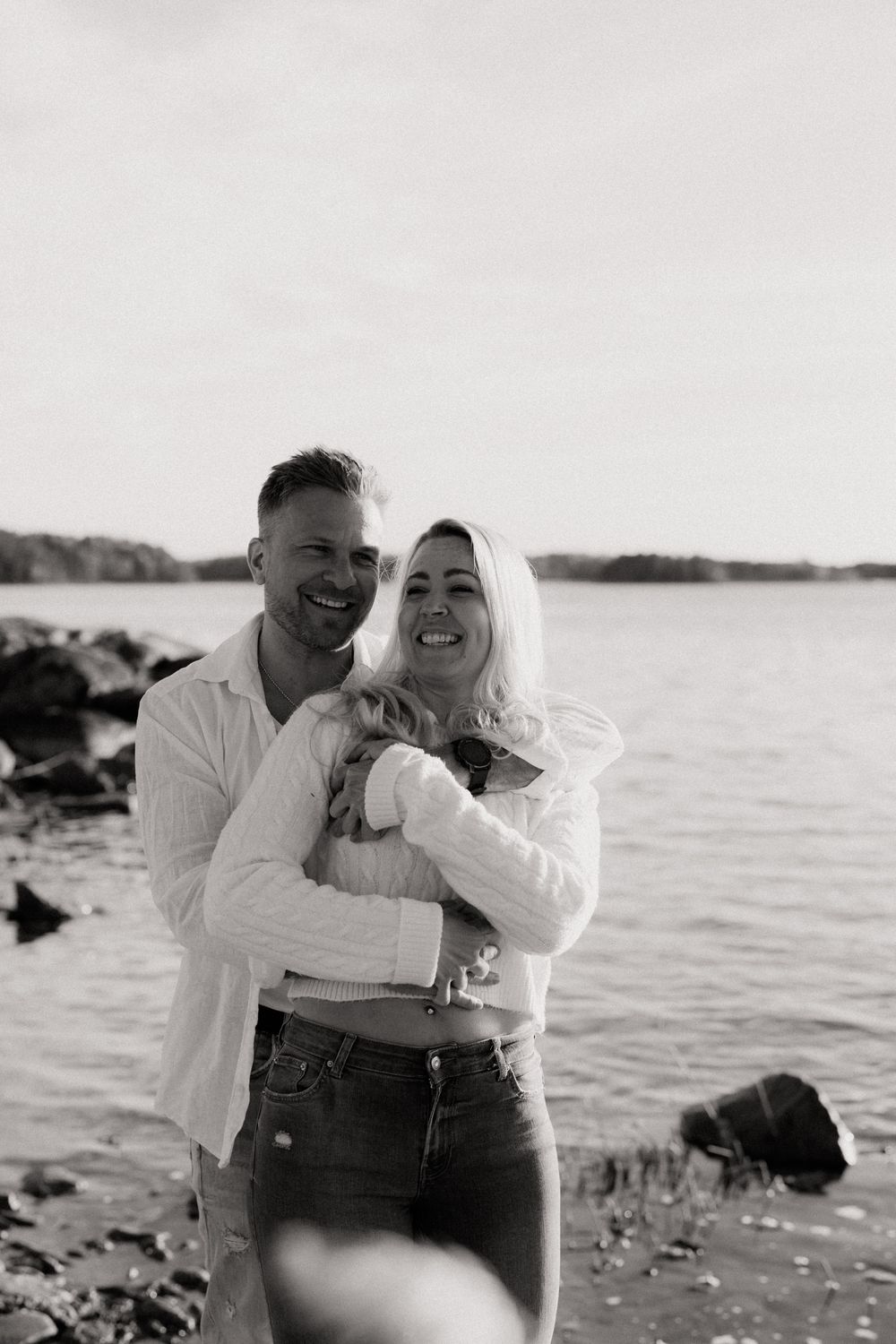 Black and white photo of a smiling couple embracing on a rocky beach with water and trees in the background.